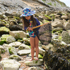 A child wearing a UV Protective Sun Hat Multi Check stands on rocks at the beach, holding a net and looking downwards.